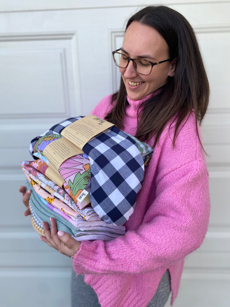 Pictured is Ashleigh, the Owner of Love Bree in a Bright Pink Knitted Jumper holding a pile of products including Heat Packs and Burp Cloths, Ashleigh has Brown Hair and Glasses and is looking down towards the pile and smiling.